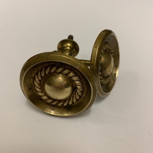 May include: Two antique brass cabinet knobs with a rope design around the edge. The knobs have a raised center and are attached to a metal post.