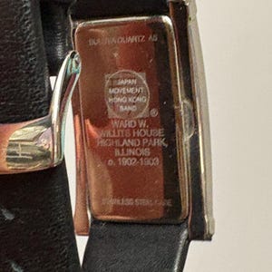 May include: A rectangular wristwatch with a black leather band. The watch face is silver-toned, and the back of the watch has text that reads "INAVA QUARTZ AS" and "JAPAN MOVEMENT HONG KONG BAND". The watch is a vintage item.