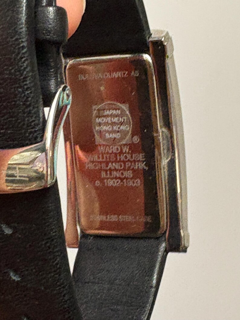 May include: A rectangular wristwatch with a black leather band. The watch face is silver-toned, and the back of the watch has text that reads "INAVA QUARTZ AS" and "JAPAN MOVEMENT HONG KONG BAND". The watch is a vintage item.