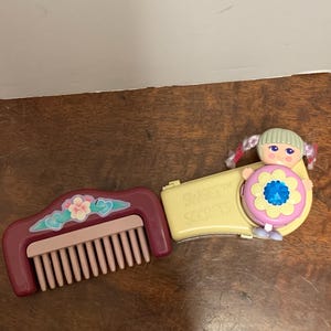 May include: A vintage toy set featuring a burgundy comb with a floral design and a yellow compact with a doll figure. The compact has the text "Sweet Secrets" and a flower-shaped design with a blue gem.