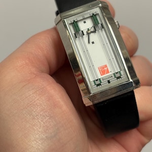 May include: A rectangular Bulova watch with a silver case and a white face featuring green and black geometric designs. The watch has a black leather band and the Bulova logo is visible. The watch is held in a hand.