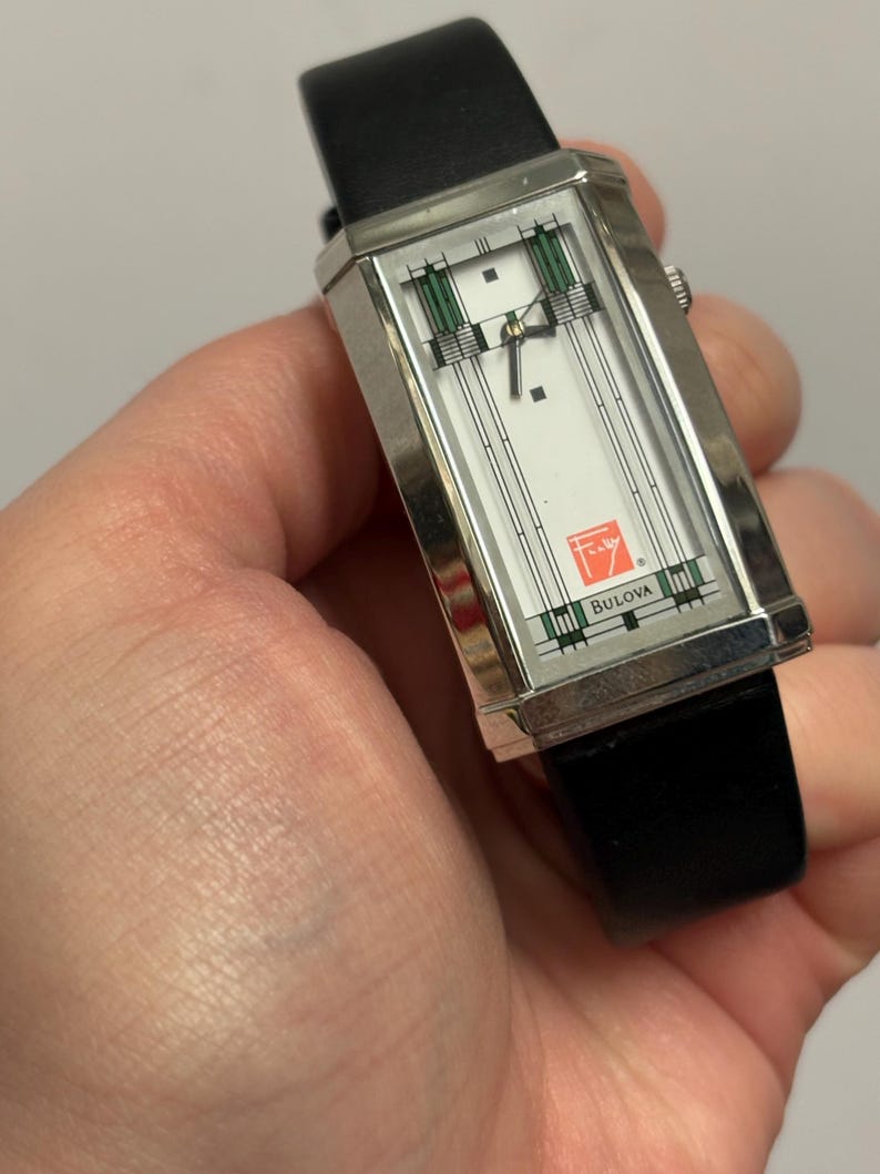 May include: A rectangular Bulova watch with a silver case and a white face featuring green and black geometric designs. The watch has a black leather band and the Bulova logo is visible. The watch is held in a hand.