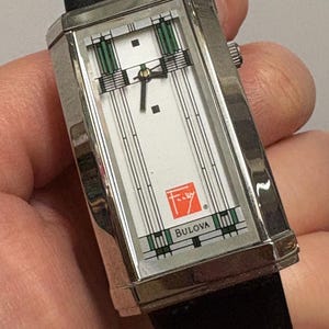 May include: A rectangular Bulova wristwatch with a silver-tone case and a black leather band. The watch face is white with green and black geometric designs. The Bulova logo and a red emblem are visible.