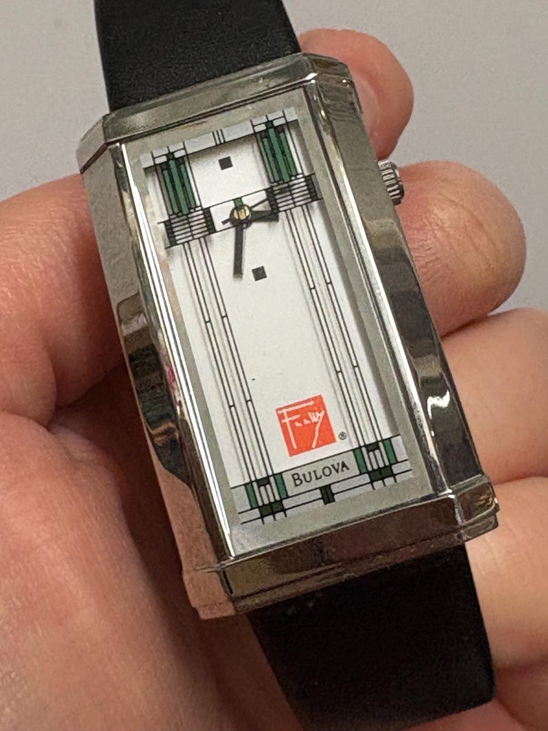 May include: A rectangular Bulova wristwatch with a silver-tone case and a black leather band. The watch face is white with green and black geometric designs. The Bulova logo and a red emblem are visible.