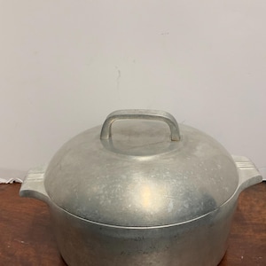 May include: A silver aluminum pot with a lid and a handle. The pot has a rounded bottom and a slightly flared rim. The lid has a knob on top and a small lip around the edge.