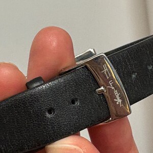 May include: Close-up of a black leather watch strap with a silver buckle. The buckle has a signature engraved on it. The strap is secured with a silver clasp.