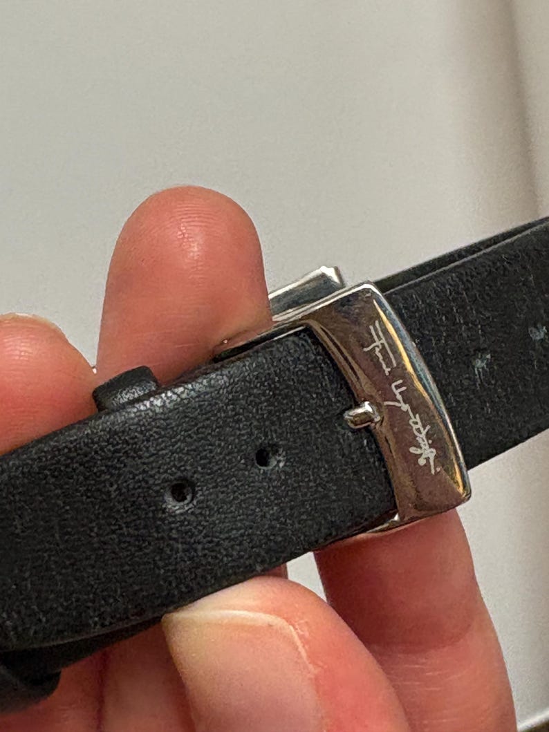 May include: Close-up of a black leather watch strap with a silver buckle. The buckle has a signature engraved on it. The strap is secured with a silver clasp.