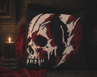May include: A black decorative pillow with a cross-stitch design of a skull in red and off-white. The skull design is centered on the pillow, with the background a solid black. A lit candle and red fabric are in the background.