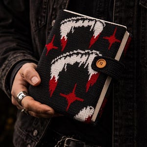 May include: Black notebook with a knitted cover featuring a repeating pattern of white fangs with red accents and red starbursts. The notebook has a black strap with a wooden button closure. The edges of the pages are visible.