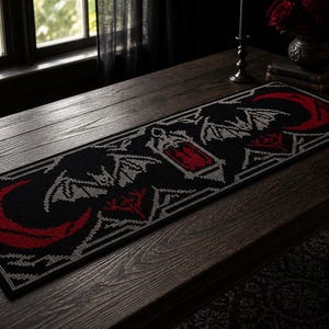 May include: A black and red table runner with a gothic design. The runner has a repeating pattern of bats, crescent moons, and geometric shapes. The design is in red and grey on a black background. The runner is on a dark wooden table.