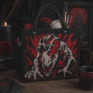 May include: Black tote bag with a cross-stitch design featuring a monster in red and gray. The bag has black handles and is filled with yarn in various colors. Crochet hooks and scissors are visible.