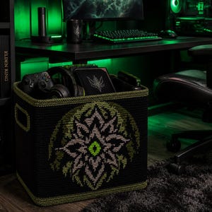 May include: A black square crochet storage basket featuring a green and grey geometric star-shaped emblem on the front. The basket has integrated side handles and a green trim along the top edge, containing a game controller and a black notebook.