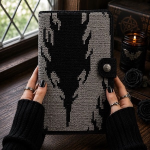 May include: A rectangular notebook cover featuring a two-tone crochet pattern in black and grey. The design displays a central jagged, flame-like black motif against a textured grey background. A side tab with a circular metal snap button secures it.