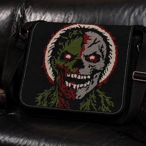 May include: A black messenger bag featuring a large, detailed cross-stitch embroidery of a split-faced zombie character. The design displays one side in green and the other in grey, with glowing red eyes, a bared-teeth grin, and a circular border.