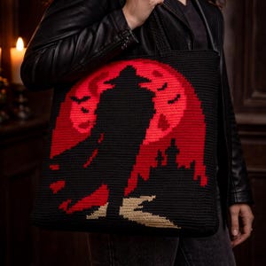 Puede incluir: Bolso tote negro con la silueta de una figura con sombrero y capa contra una luna roja. El diseño incluye murciélagos y un castillo. La bolsa está hecha de material tejido y tiene asas negras.
