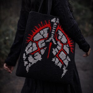Puede incluir: Bolso tote negro con un diseño de pulmón de ganchillo en gris y rojo. El bolso tiene forma rectangular con un asa. El fondo está borroso, lo que sugiere un entorno al aire libre.