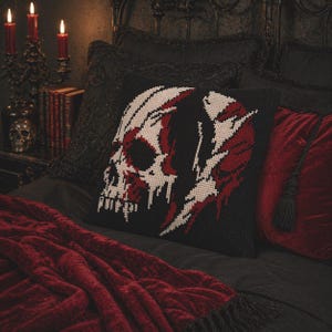 Könnte beinhalten: Ein schwarzes Zierkissen mit einem Kreuzstich-Schädeldesign in Weiß und Rot. Das Kissen liegt auf einem Bett mit einer roten Decke und anderen schwarzen und roten Kissen. Ein Kerzenleuchter und Bücher sind im Hintergrund.