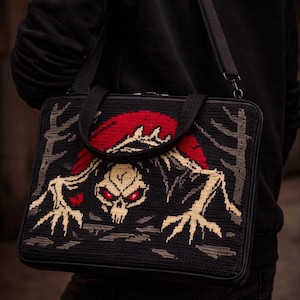 May include: Black laptop bag with a detailed, embroidered design. The design features a skull with red eyes and a red arched shape above it, with tan and gray accents. The bag has a black shoulder strap and a handle.