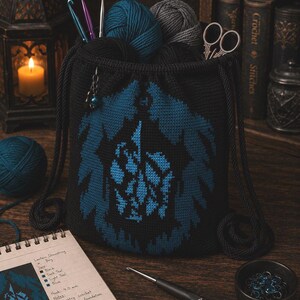 Puede incluir: Una bolsa negra de ganchillo con un diseño azul y negro, llena de hilo, ganchos de ganchillo y tijeras. También se ve un cuaderno con instrucciones de ganchillo y una vela encendida en una linterna. El tamaño del ganchillo es de 4,0 mm.
