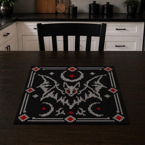 Può includere: Un sottopentola nero con un disegno di pipistrello. Il pipistrello è grigio con accenti rossi e il bordo è bianco e rosso. Il sottopentola è su un tavolo di legno marrone scuro. La cucina sullo sfondo ha mobili bianchi e un piano di lavoro nero.