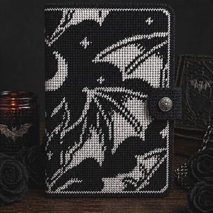 May include: Black and white cross-stitched book cover featuring a bat and moon design. The cover has a button closure and is surrounded by black roses and a candle. The overall theme is gothic.