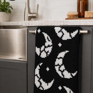 May include: Black and white decorative kitchen towel featuring a repeating pattern of crescent moons and stars. The towel is hanging on a silver bar in a kitchen setting. The design is a pixelated style.