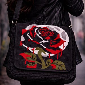 Könnte beinhalten: Schwarze Umhängetasche mit einem großen, gestickten Rosendesign. Die Rose hat rote Blütenblätter, grüne Blätter und einen grauen Mond im Hintergrund. Die Tasche hat einen schwarzen Riemen und einen Klappenverschluss.