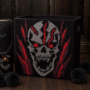 May include: A square black storage basket featuring a crocheted graphic of a grey skull with glowing red eyes and jagged red lightning-like accents. The basket has a textured surface and a wide open top, showcasing a detailed needlework design.