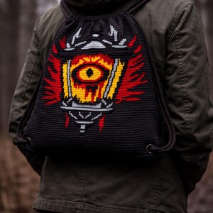 Puede incluir: Mochila negra con cordón y un diseño vibrante y pixelado de un ojo estilizado en amarillo, rojo y blanco. La mochila se lleva sobre una chaqueta verde, con los cordones visibles. El diseño de la bolsa recuerda a un tema de fantasía o juegos.