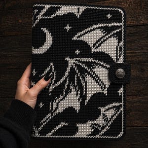 May include: Black and white cross-stitch tablet cover with a bat and crescent moon design. It has a button closure and a decorative button. The cover is made of a textured material.