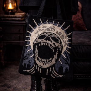 Può includere: Borsa tote nera con un design a teschio. Il teschio è al centro, circondato da un motivo a raggiera. La borsa è tenuta da una persona che indossa stivali neri e una giacca nera. Il teschio è nero e beige.