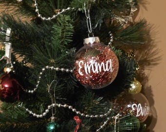 May include: A festive Christmas tree adorned with various decorations. Clear baubles filled with red glitter and the names "Emma" and "Elvis" in white lettering are visible. Other ornaments include green, gold, and red balls, and silver beaded garland.