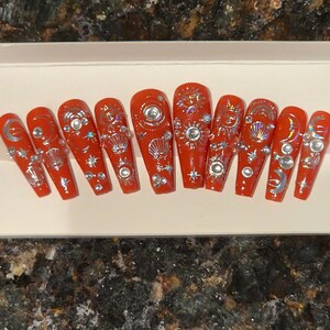 May include: Set of ten red press-on nails with a celestial theme. The nails are decorated with silver glitter, moons, stars, and suns.