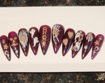May include: Set of ten burgundy stiletto false nails with white and gold Halloween themed designs. The designs include ghosts, hearts, spiderwebs, and the word "BOO".