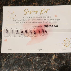 Op de afbeelding: Press-on nagel maatset met tien doorzichtige kunstnagels genummerd van 0 tot 9. De set is voor amandelvormige nagels. De tekst "Sizing Kit" is geschreven in zwarte cursieve letters. De tekst "FOR PRESS-ON NAILS" is geschreven in zwarte letters. De tekst "Use the nails to find your perfect fit for each finger. Write down your sizes from thumb to pinky." is geschreven in zwarte letters. De tekst "NAIL SHAPE Almond" is geschreven in zwarte letters.