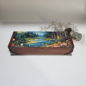 May include: A rectangular wooden box with a forest scene on the lid. The box is brown with metal corner protectors. The lid features a colorful painting of a river flowing through a forest with trees and flowers.