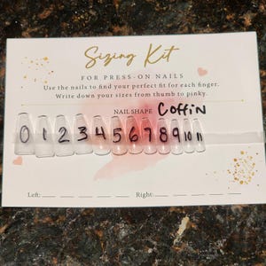 Pode incluir: Um kit de dimensionamento para unhas postiças. O kit inclui amostras de unhas transparentes em forma de caixão, numeradas de 0 a 11. O cartão fornece instruções para medir os tamanhos das unhas, do polegar ao mindinho. O texto "Sizing Kit" é dourado.