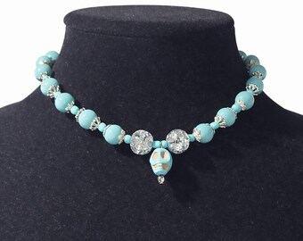 Puede incluir: Un collar de cuentas turquesas con detalles plateados y un colgante de calavera. El collar presenta cuentas grandes y redondas de color turquesa, intercaladas con cuentas de filigrana plateadas. El colgante es una pequeña calavera turquesa.