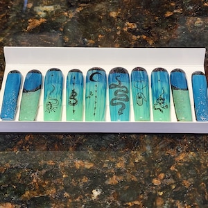 Op de afbeelding: Set van tien blauwe en groene ombre press-on nagels met verschillende zwart-witte ontwerpen, waaronder slangen, manen en harten.