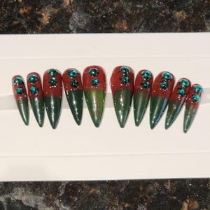 Puede incluir: Conjunto de diez uñas postizas largas y puntiagudas con un diseño degradado rojo y verde. Cada uña tiene tres pedrerías color turquesa.