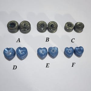 May include: An assortment of six pairs of ear gauges. The top row features circular gauges with sun and moon designs in gray. The bottom row shows heart-shaped gauges in blue, also with sun and moon designs. The gauges are displayed on a white surface.