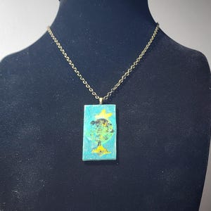 May include: A pendant necklace featuring a rectangular pendant with a teal background and a globe design. The globe is green and black with yellow accents. The chain is gold-coloured.