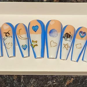 May include: Set of ten blue and white false nails with various zodiac and celestial designs, including a crescent moon, a star, a heart, a bull, and the word "Taurus".