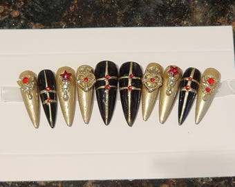May include: Set of ten false nails with a gold and black design. Each nail has a different design, including a black and gold stripe pattern, a gold heart with red jewels, and a gold star with red jewels.
