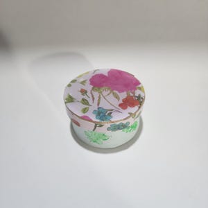 May include: A round, floral-patterned box with a lid. The lid features a large pink rose and other colorful flowers on a white background. The box's sides are light green with floral accents.