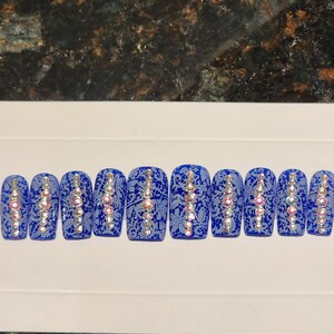May include: A set of ten blue press-on nails with a lace pattern and a vertical line of rhinestones. The nails are arranged in a row on a white surface.