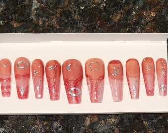 May include: Set of ten long, almond-shaped, false nails with a clear base and a red glitter gradient. Each nail has a different silver design, including moons, stars, and circles.