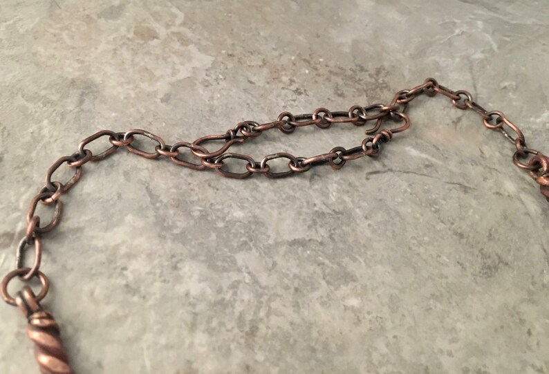 May include: A close-up of a copper chain necklace with a twisted copper pendant. The chain is made of oval links.