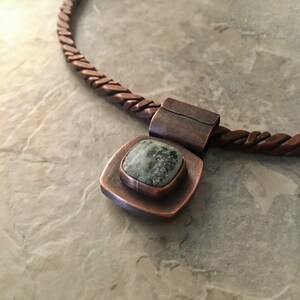 May include: A copper necklace with a braided chain and a square pendant featuring a green stone.
