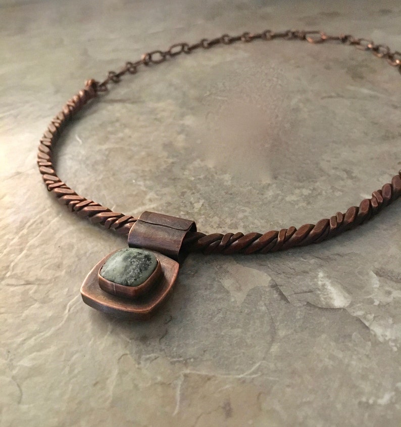 May include: A copper necklace with a braided chain and a square pendant featuring a green stone.